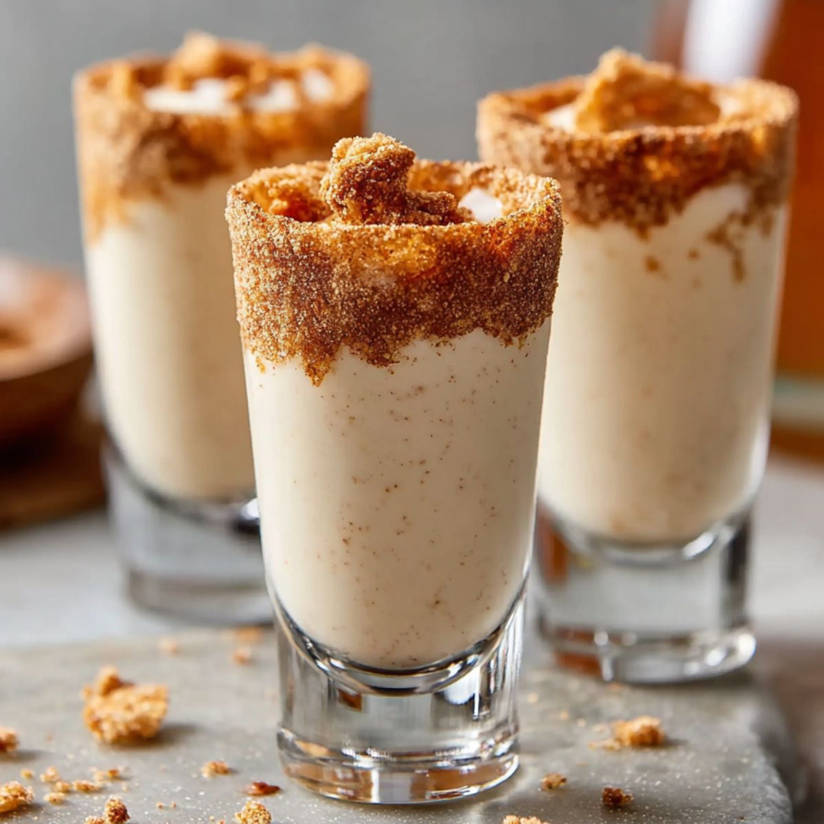 Cinnamon Toast Crunch Shots in clear glass shot glasses, filled with creamy cinnamon liqueur and rimmed with crushed cereal. The drinks sit on a light gray surface sprinkled with crumbs and a few sugar-coated cubes, creating a cozy, real-kitchen vibe.
