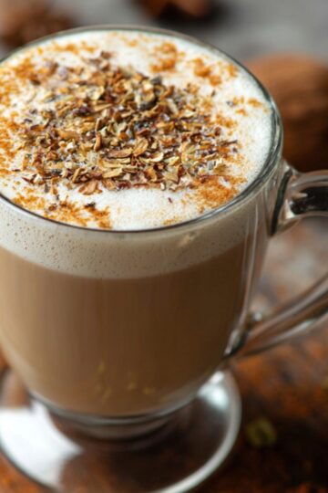 “Glass mug filled with creamy Coconut Chai topped with frothed milk, sprinkled with cinnamon and crushed spices on a rustic wooden surface.”