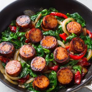 Sliced smoked sausage pieces browned and served in a skillet with sautéed spinach, red bell peppers, and onions.