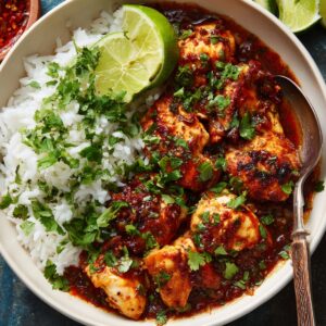 A bowl of spicy chicken in a rich red sauce served with white rice and garnished with fresh chopped cilantro and a lime wedge.