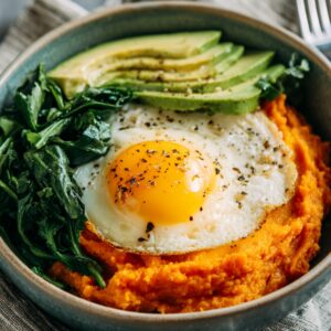 A breakfast bowl with mashed sweet potatoes, sautéed spinach, sliced avocado, and a sunny-side-up egg seasoned with pepper.