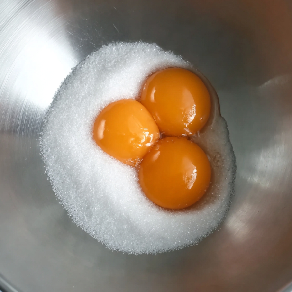 Top-down view of three bright orange egg yolks resting against a mound of white granulated sugar inside a stainless steel mixing bowl.