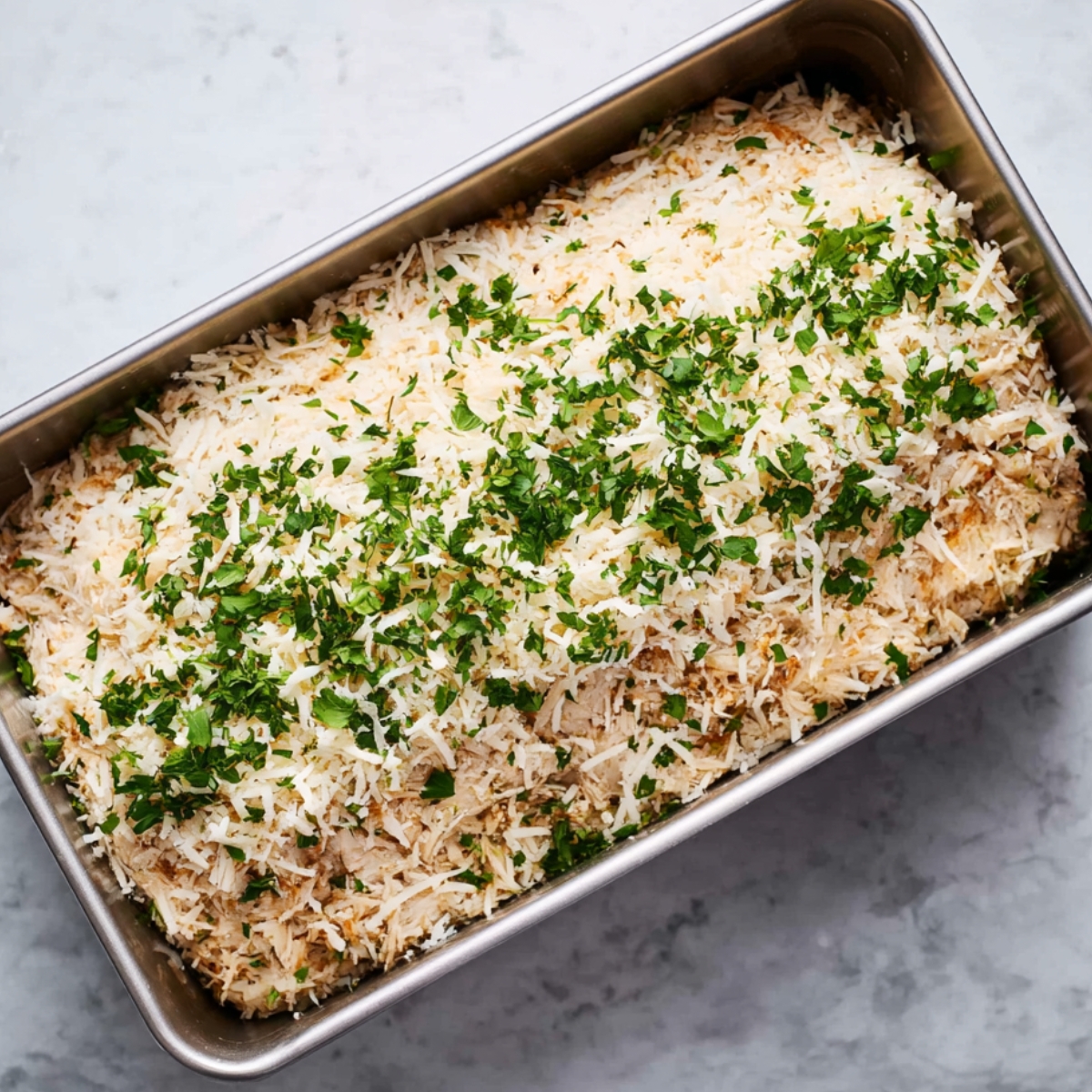 Top-down view of baked garlic parmesan chicken meatloaf in a loaf pan, golden brown with melted cheese and parsley garnish, surrounded by garlic cloves and cheese