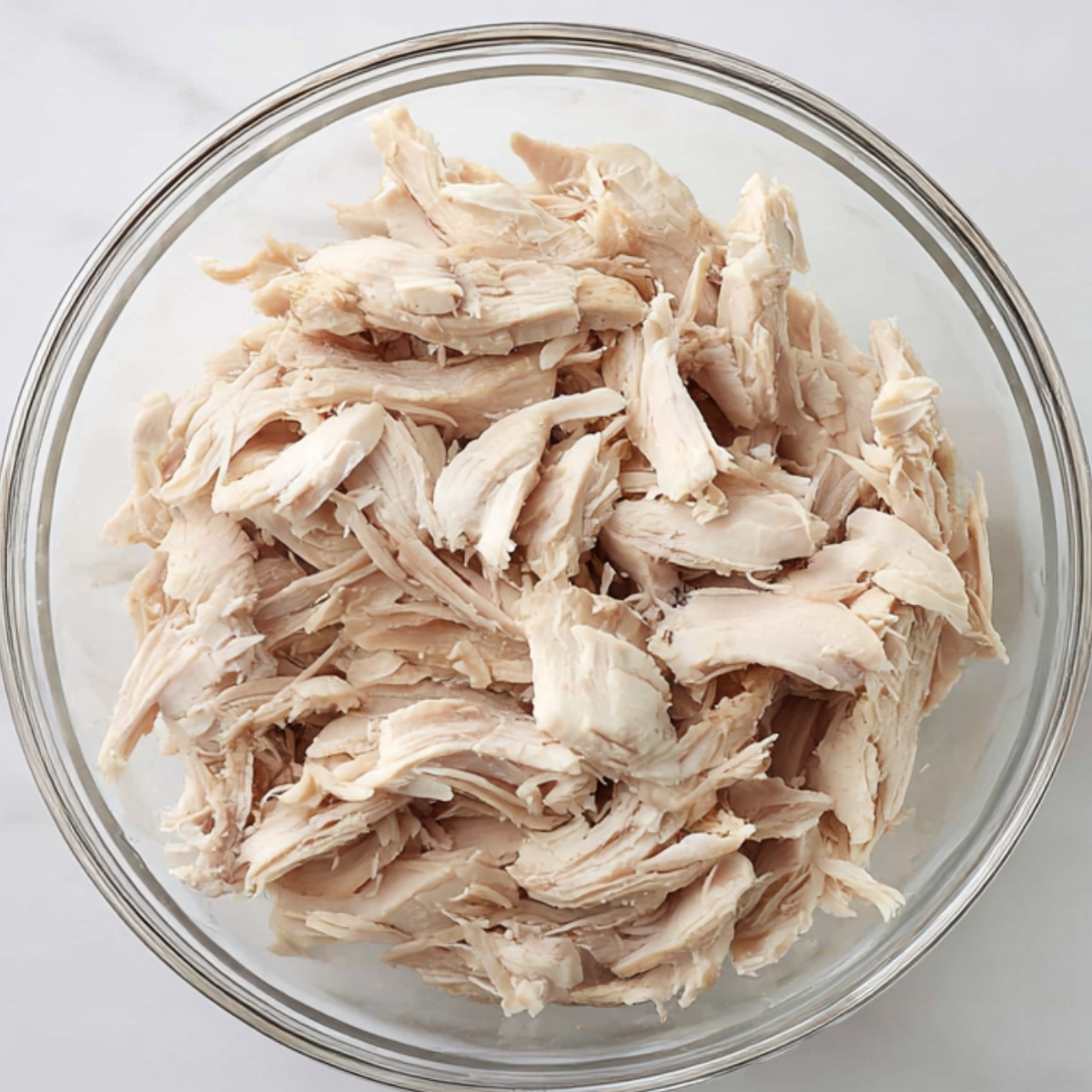 Top-down view of a clear glass bowl filled with plain shredded cooked chicken on a white marble countertop.