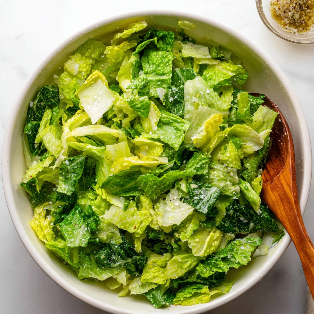 A fresh bowl of chopped romaine lettuce, possibly with some seasoning or dressing on top. There is a wooden spoon beside the salad.