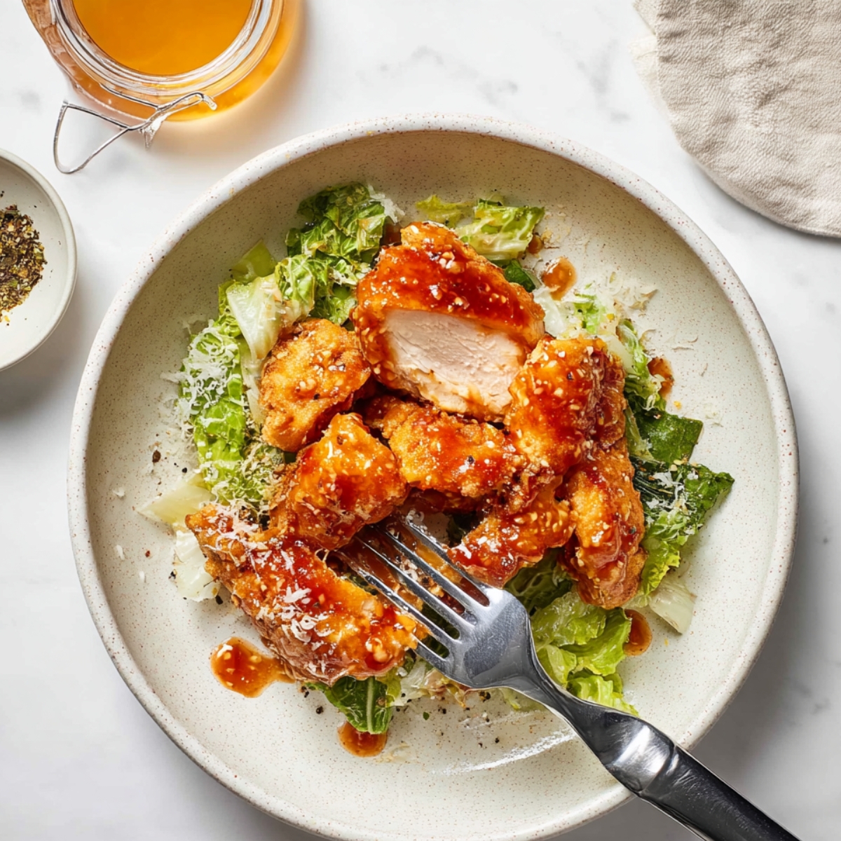 A serving of crispy, hot honey-glazed chicken pieces on top of romaine lettuce, with some Parmesan cheese sprinkled over the chicken and a fork in the foreground.