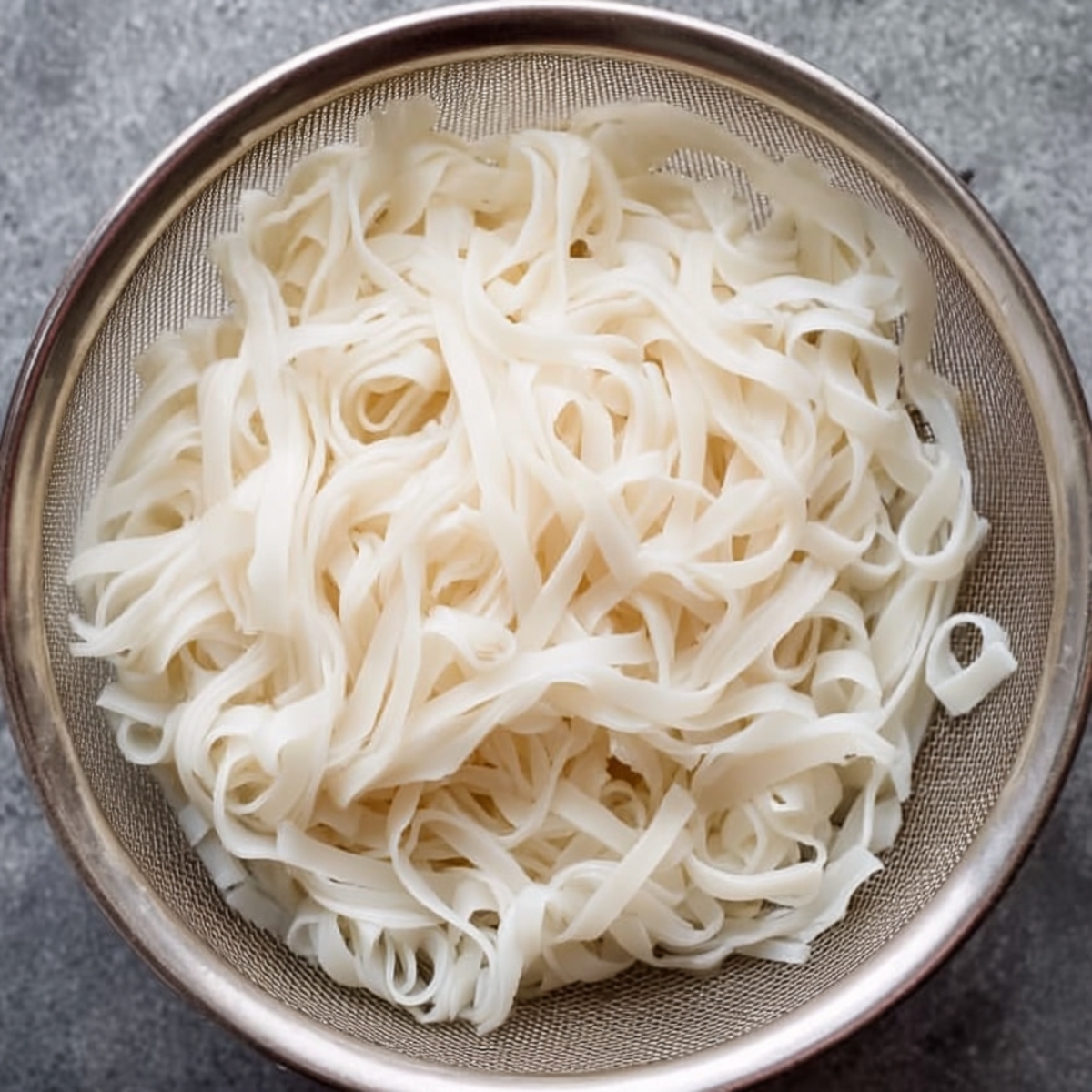 Top-down view of wide flat rice noodles draining in a fine metal strainer, pale white noodles softly curled and slightly translucent on a gray countertop.