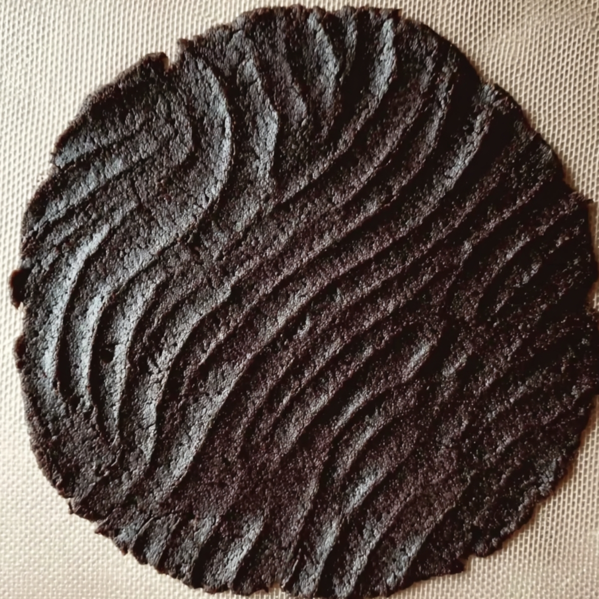 A flat, irregular round of dark chocolate dough with a rippled, textured surface resting on a light-colored baking mat.