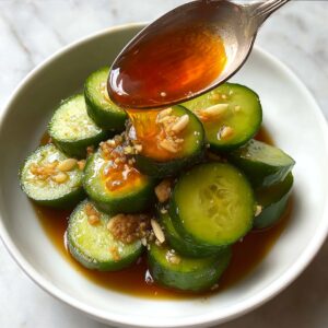 Close-up action shot of a spoon pouring glossy amber sauce over sliced cucumbers in a white bowl, highlighting garlic bits and fresh cucumber texture.