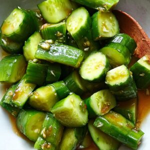 Close-up of chopped cucumbers tossed in a soy-based dressing with visible garlic pieces in a white bowl, showing glossy texture and fresh green color.