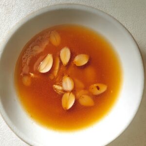 Minimalist top-down shot of an amber-colored soy and vinegar dressing with floating garlic slices in a white bowl under soft natural light.