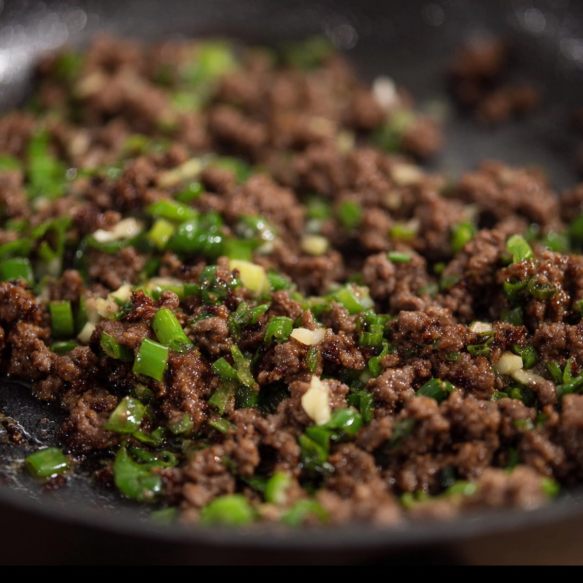 Close-up of browned ground beef sautéed with chopped green onions and garlic in a dark skillet, glistening with oil and caramelized bits.