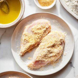 Two chicken breasts are coated with a breadcrumb or flour mixture, ready to be cooked. A bowl of oil for dipping and some other ingredients are placed beside it.