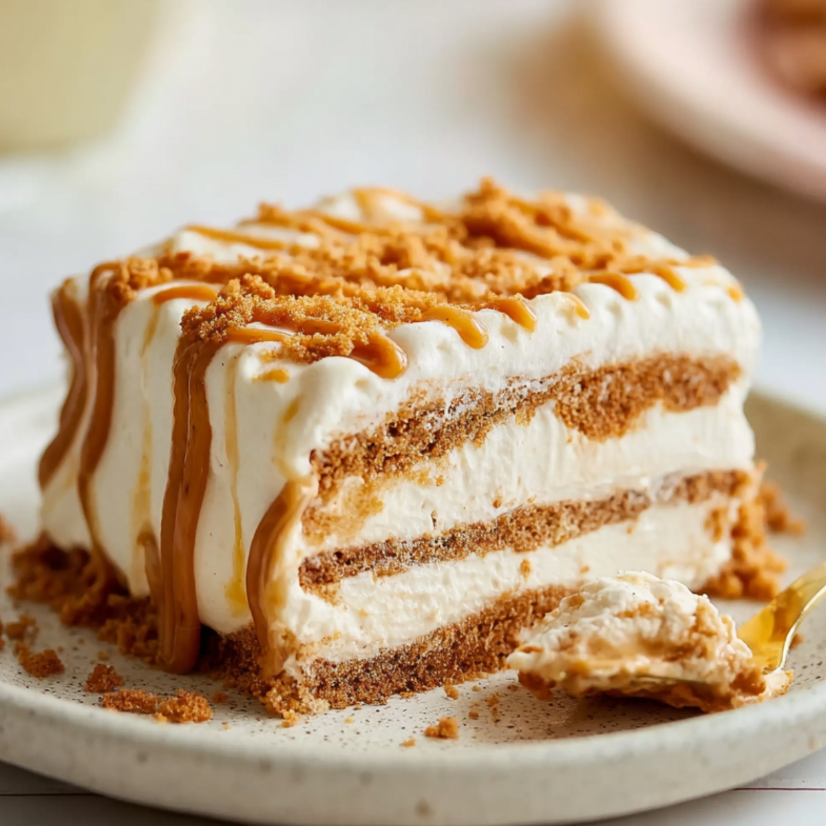 Biscoff Icebox Cake Slice of Biscoff icebox cake with layers of creamy vanilla filling and Biscoff cookies, topped with cookie crumbs and caramel drizzle on a ceramic plate.