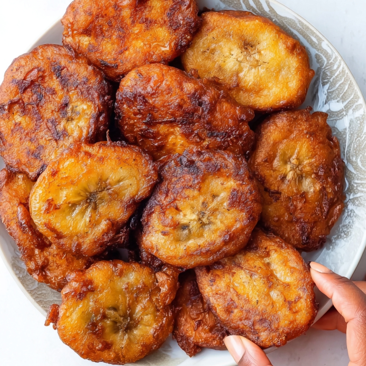 Jamaican Banana Fritters A plate filled with freshly fried banana fritters, golden and crispy, piled high for an appetizing look. A hand is reaching for one of the fritters, ready to enjoy the delicious treat. The fritters glisten with a slight sheen, showcasing their perfectly fried texture against a clean background.