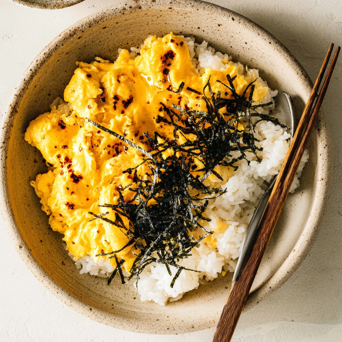 Miso scrambled eggs Overhead view of creamy miso scrambled eggs served over steamed white rice in a rustic ceramic bowl, topped with shredded nori and chili flakes.