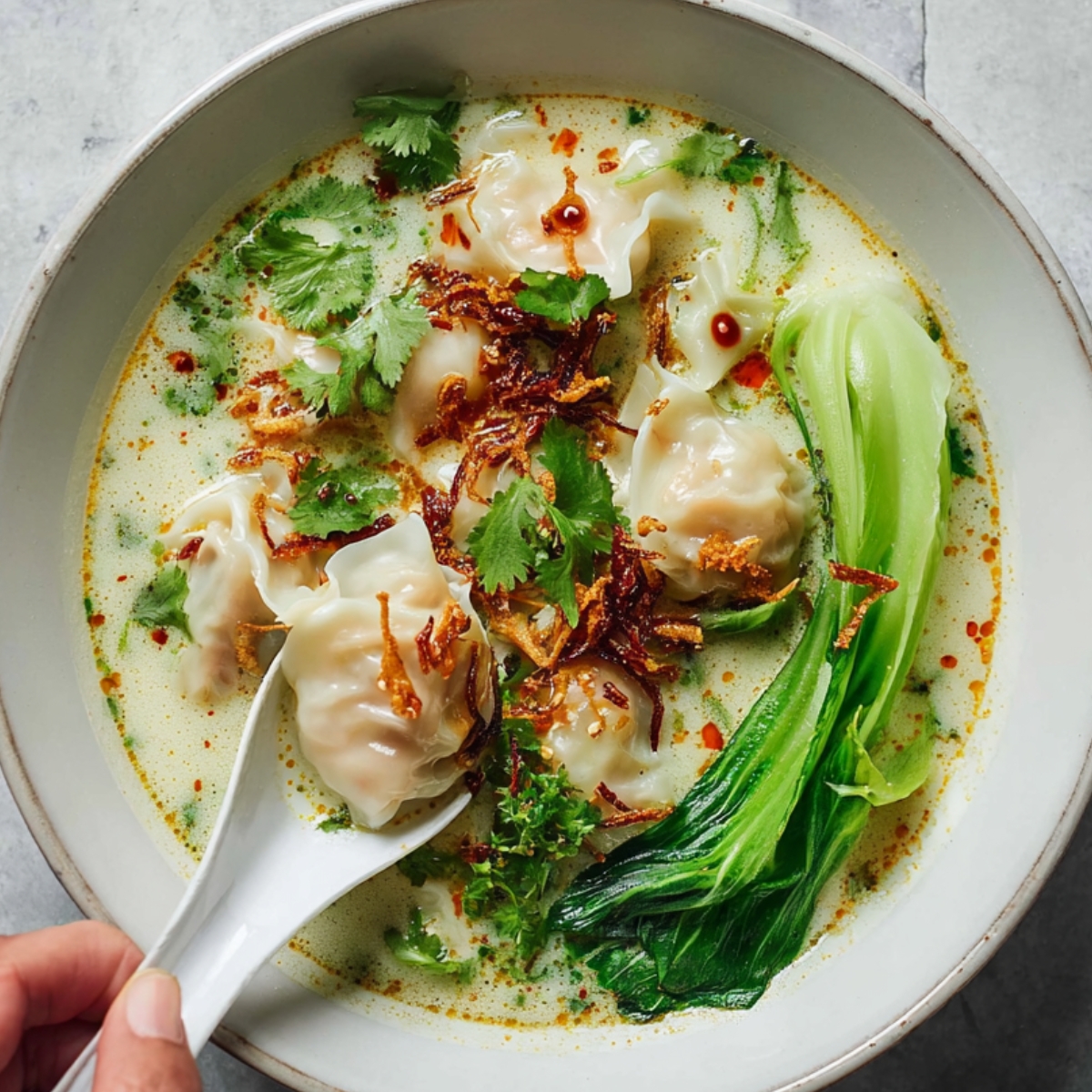 Thai Green Curry Dumpling Soup Thai green curry dumpling soup in a white bowl with shrimp dumplings, baby bok choy, cilantro, crispy fried shallots, and chili oil, with a spoon lifting a dumpling from the creamy broth.