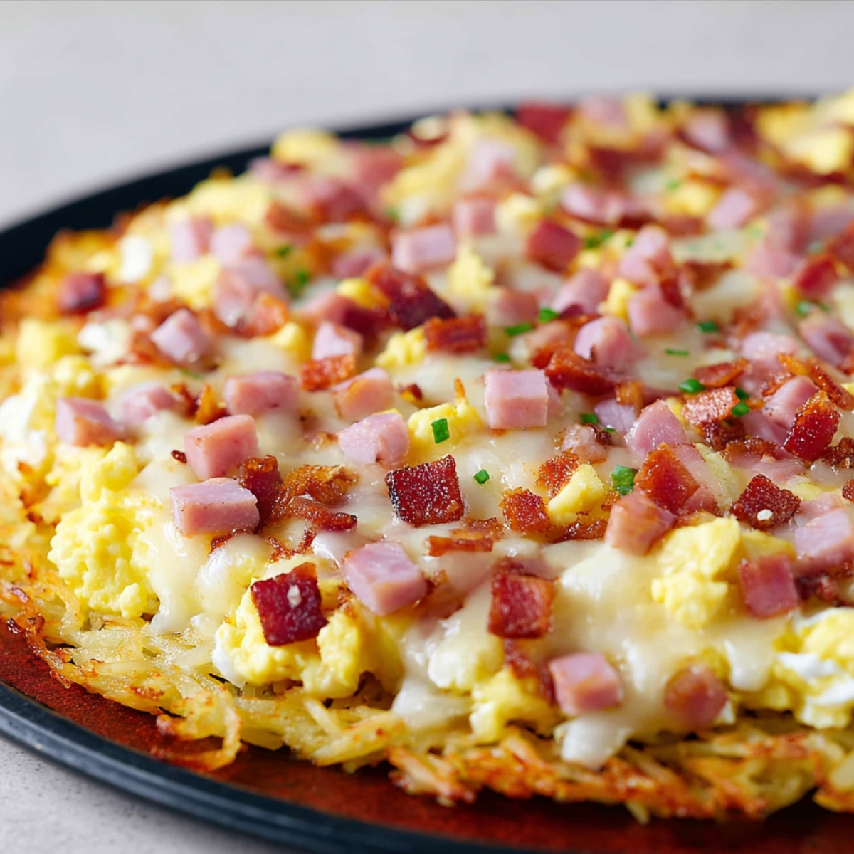 Close-up of breakfast pizza showing fluffy scrambled eggs, melted cheese, crispy bacon bits, and diced ham on a hash brown potato crust.
