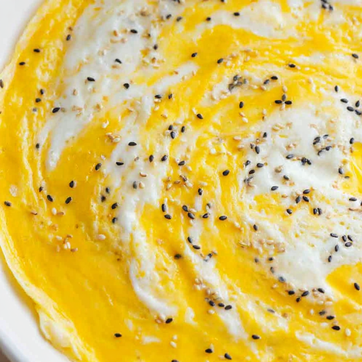 Close-up of jianbing crepe batter with swirled egg cooking on a hot surface, sprinkled with black and white sesame seeds.