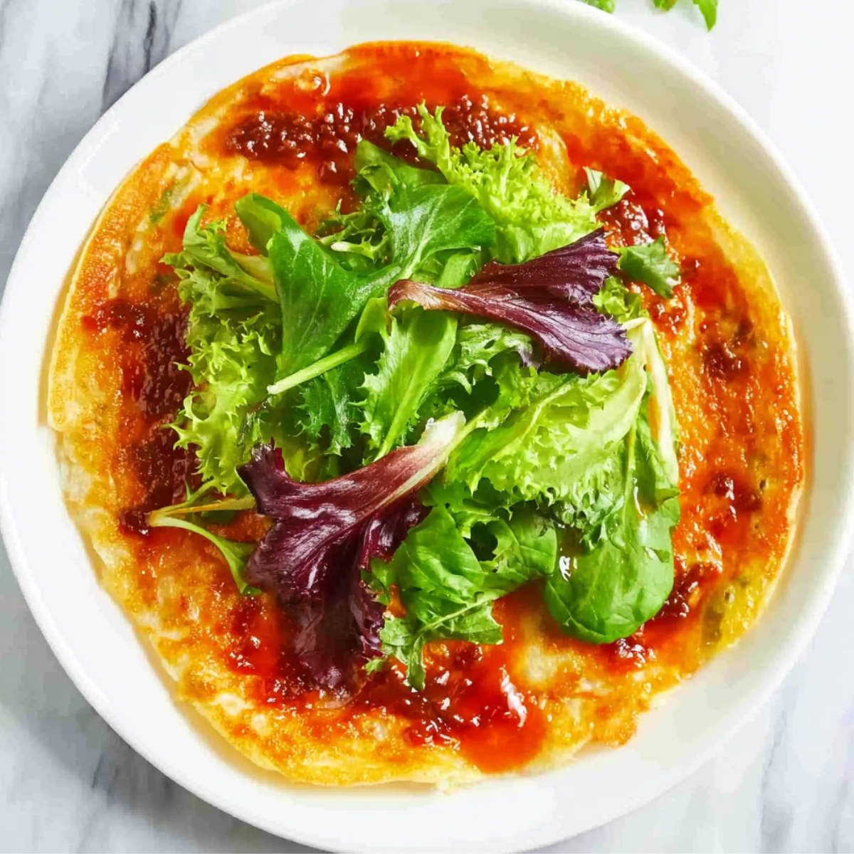 Top-down view of jianbing crepe spread with red chili sauce and topped with fresh mixed greens before folding.