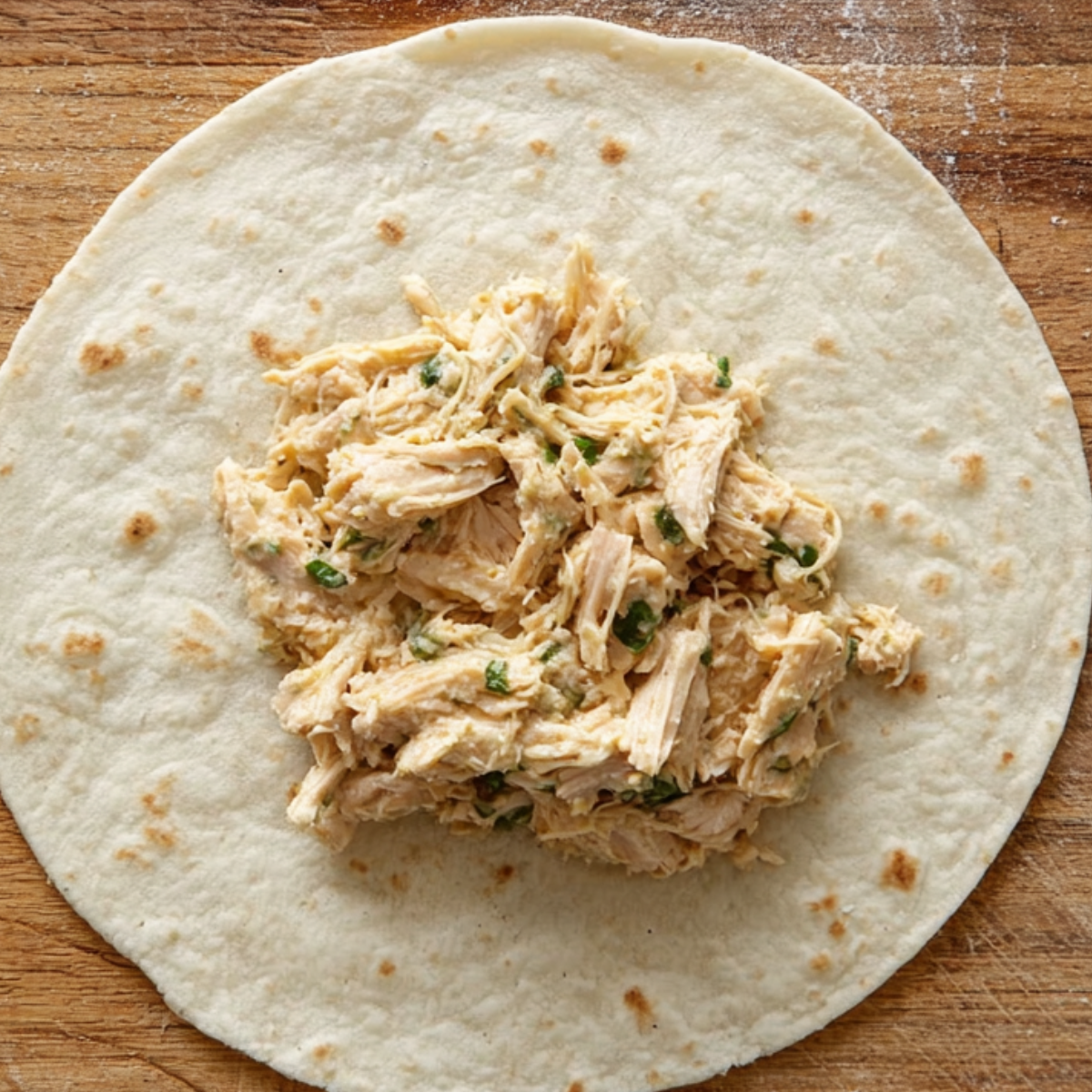 A tortilla with a scoop of seasoned shredded chicken mixture placed in the center, ready to be rolled up into taquitos.