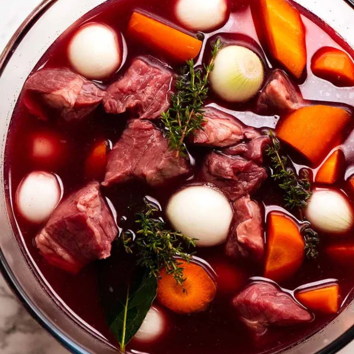 Close-up of beef chunks marinating in red wine with pearl onions, carrots, thyme sprigs, and bay leaves, all soaking in the rich, deep red marinade. The ingredients are fresh and vibrant, ready for slow cooking.