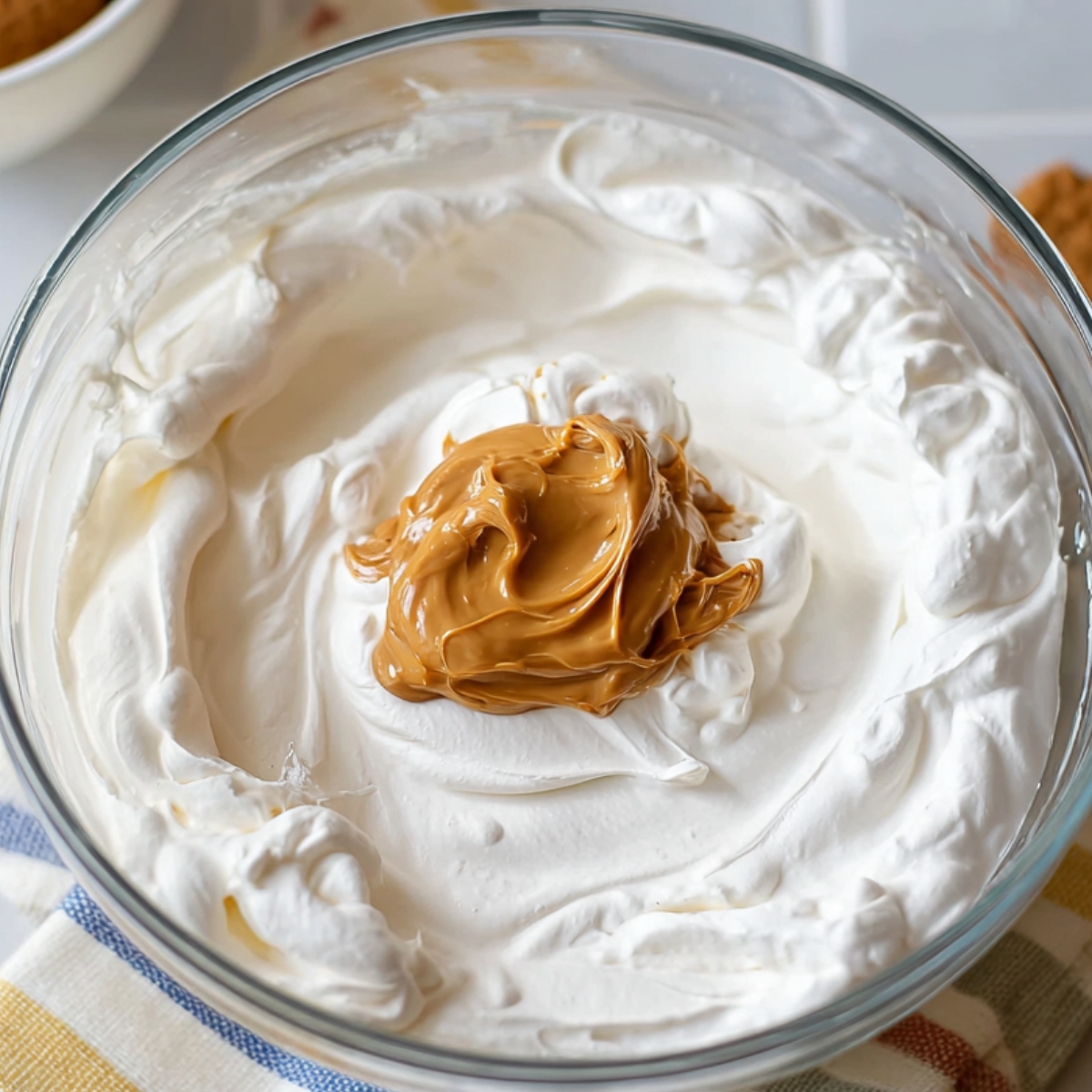 Glass mixing bowl filled with whipped cream and a dollop of Biscoff cookie butter in the center, placed on a kitchen towel with cookies nearby.