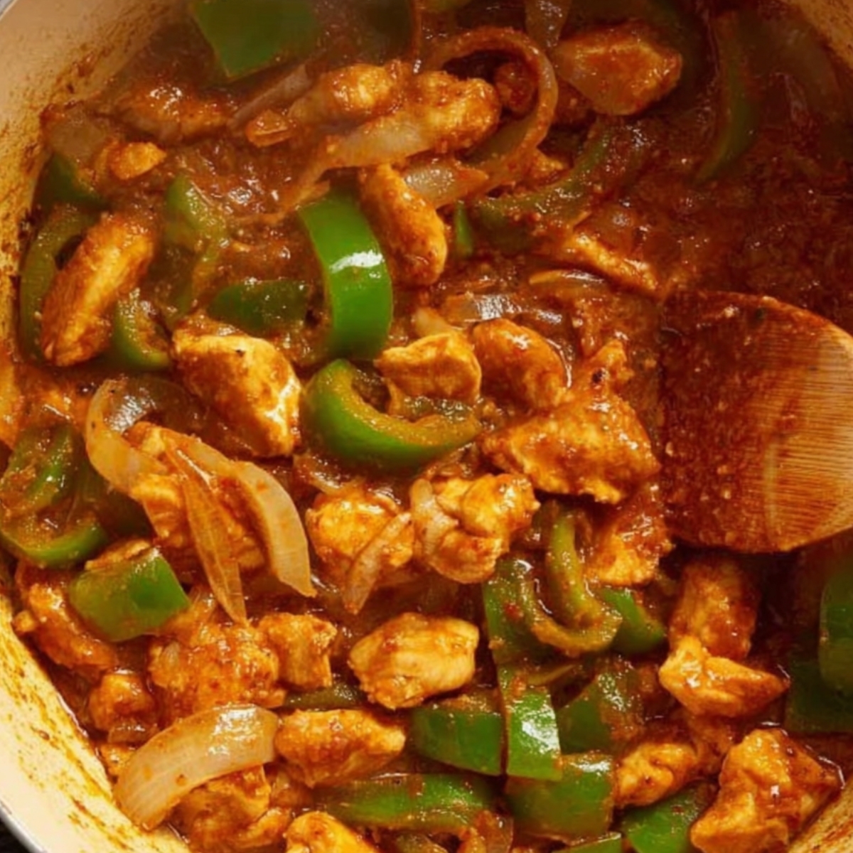 Chicken pieces cooked in a rich, spiced sauce with sliced onions and green bell peppers, simmering in a pan with a wooden spoon.