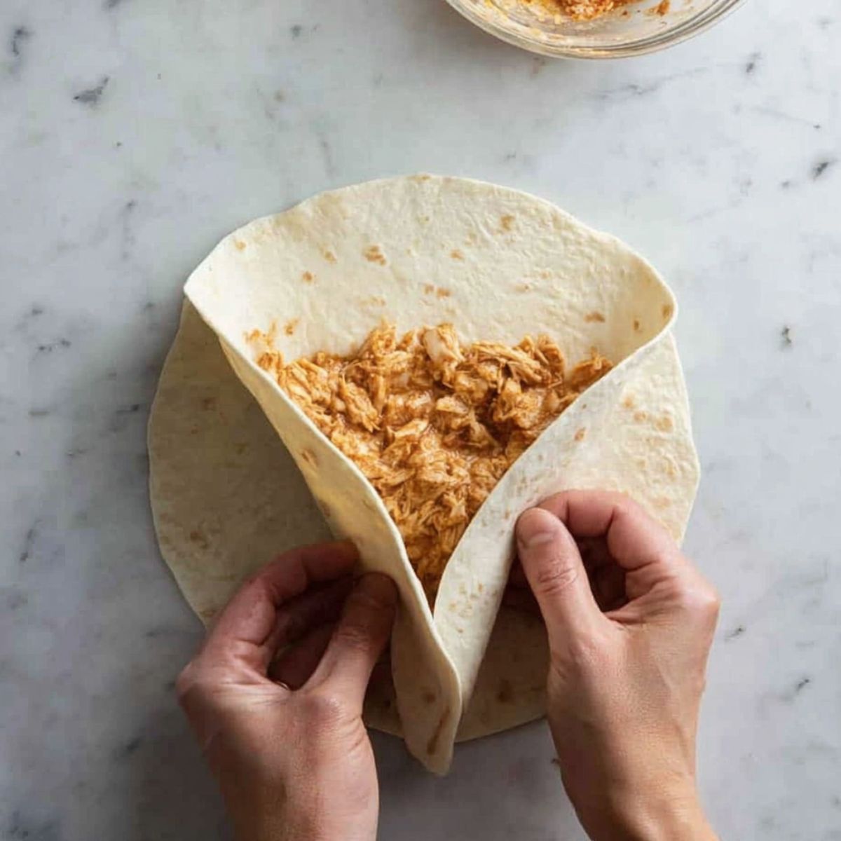 Hands folding the sides of a flour tortilla over shredded chicken filling, demonstrating how to wrap a chimichanga step by step.