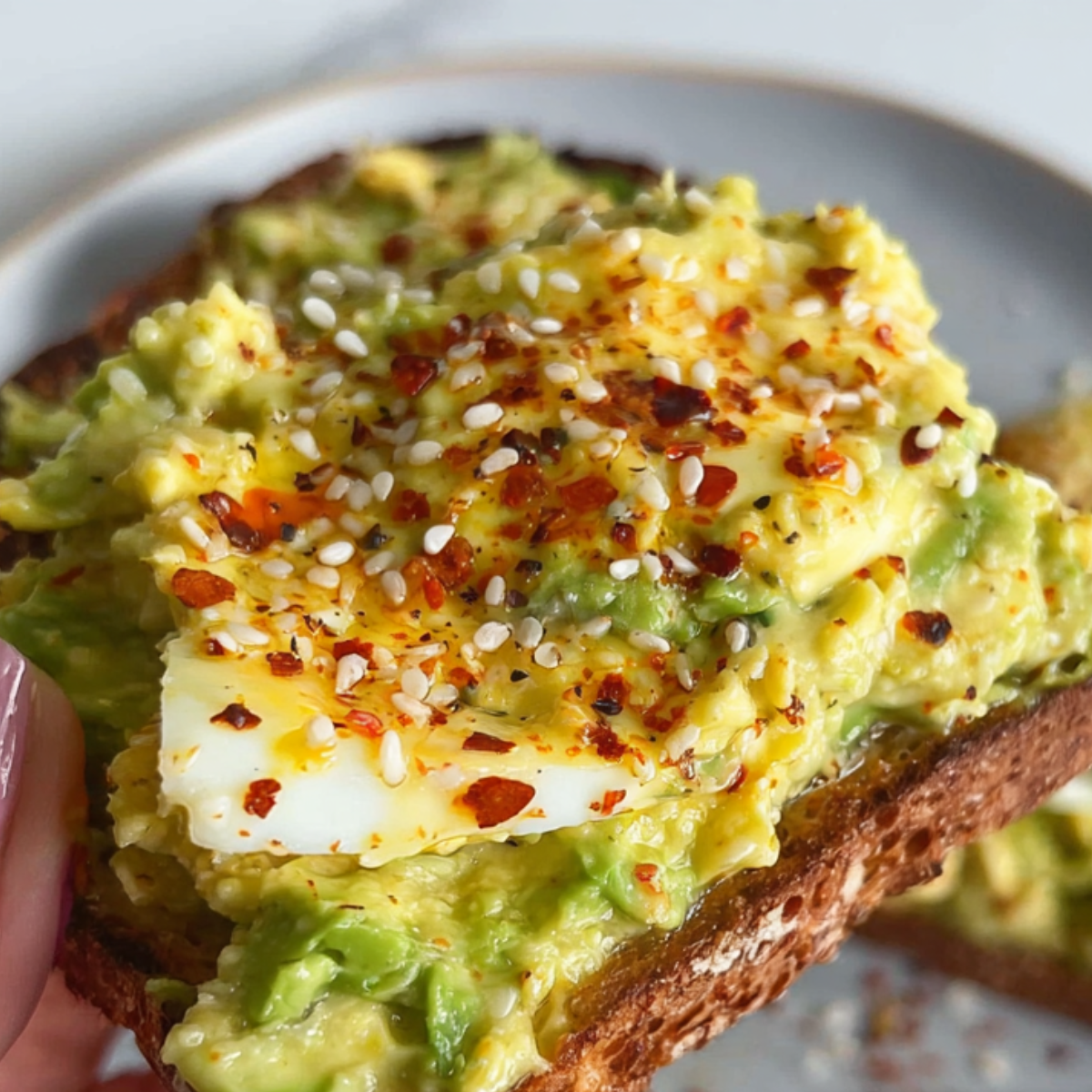A hearty slice of avocado toast topped with a soft-boiled egg, red pepper flakes, and a dash of seasoning. This simple, yet flavorful dish makes for an energizing breakfast or light lunch, packed with healthy fats and protein.