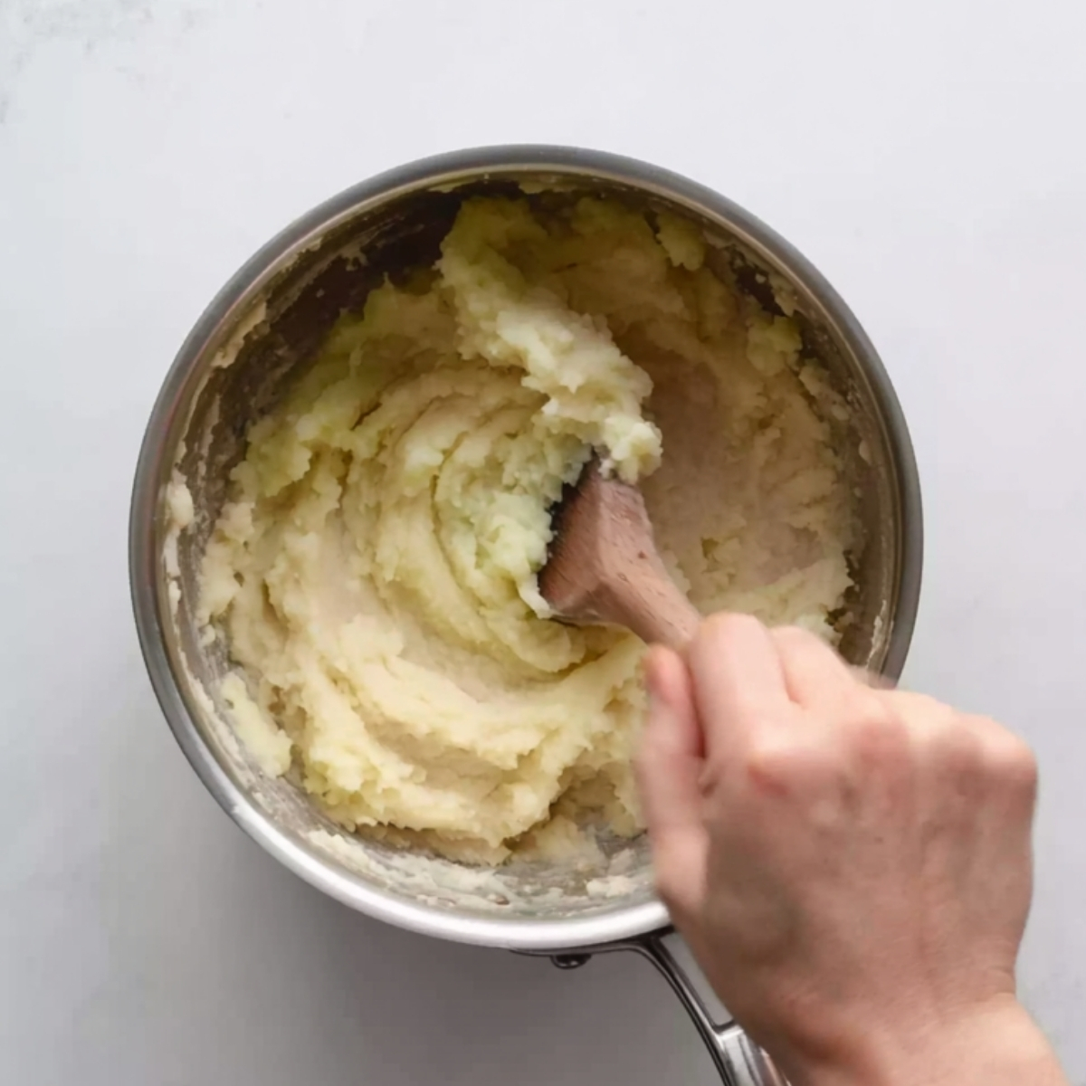 Overhead view of mashed potatoes being stirred with a wooden spoon in a stainless steel saucepan on a white countertop.