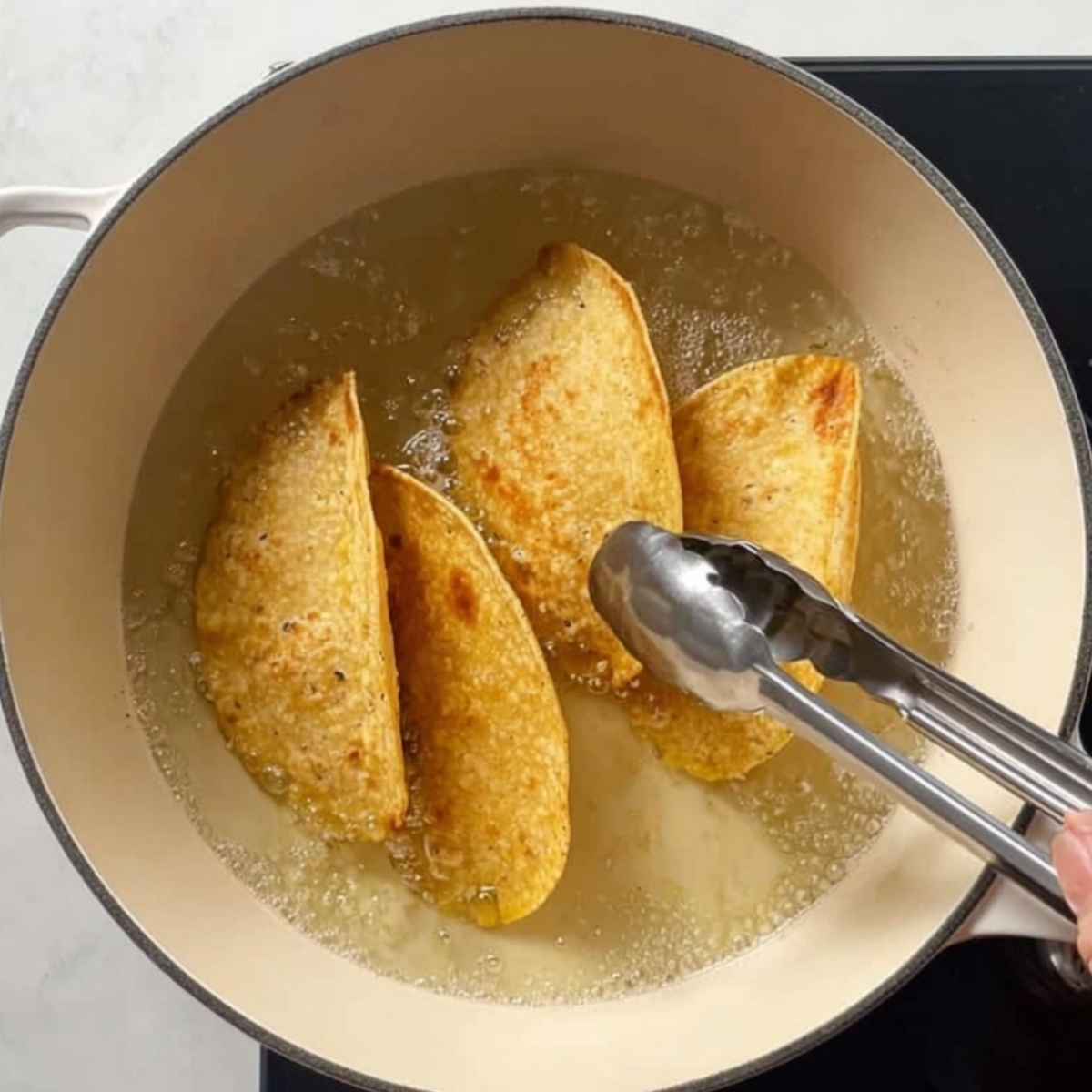 Overhead view of crispy folded corn tortillas frying in hot oil in a light-colored Dutch oven, with metal tongs flipping one taco.