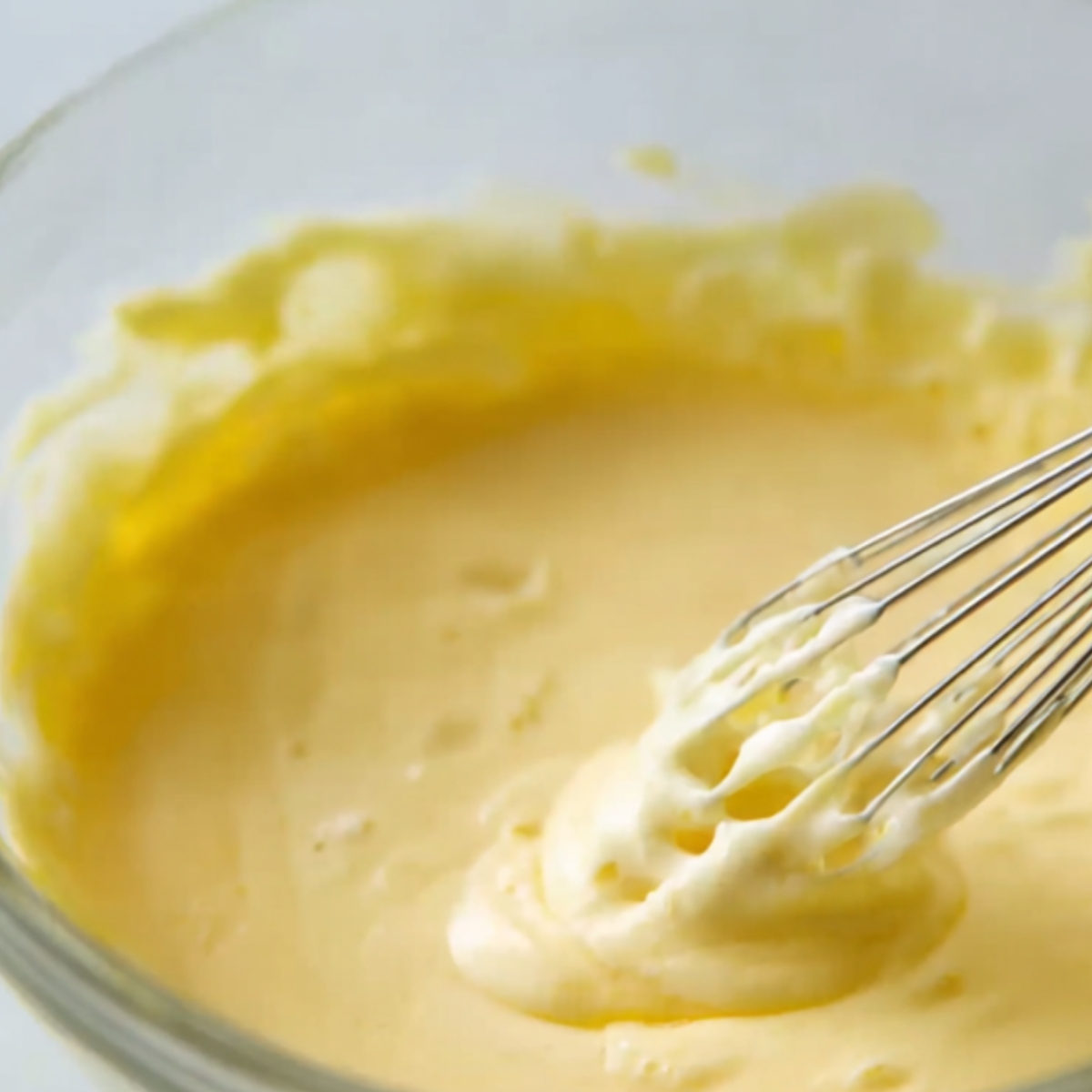 A close-up shot of a whisk being used to stir a smooth yellow mixture, likely an egg or sauce mixture, in a glass bowl.
