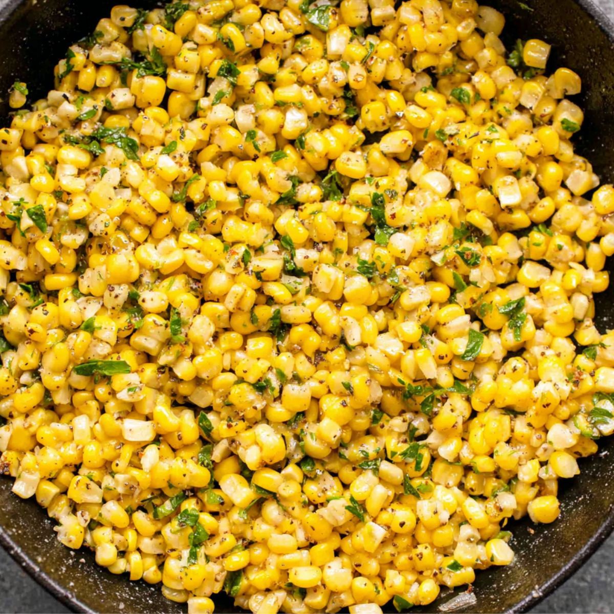 Buttery sautéed corn kernels with chopped herbs and black pepper cooked in a cast-iron skillet, photographed from above.