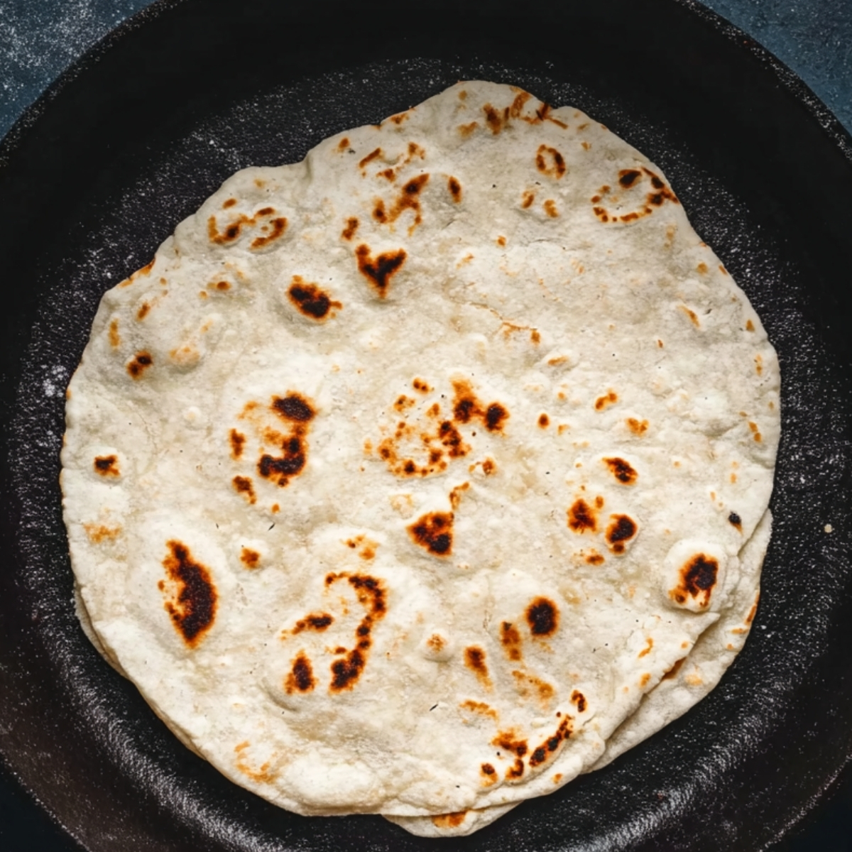 Homemade flour tortilla cooking in a cast iron skillet with golden brown char spots.