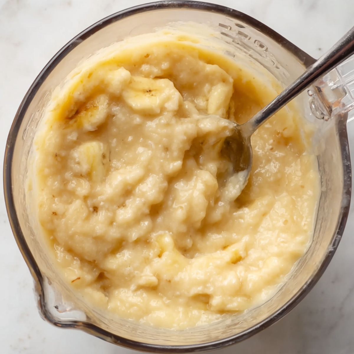 A close-up shot of a spoon stirring a thick, smooth mashed banana mixture in a glass measuring cup. The mixture is creamy with some small banana chunks, and the light shines off the smooth surface. The countertop background is clean, emphasizing the homemade feel of the preparation