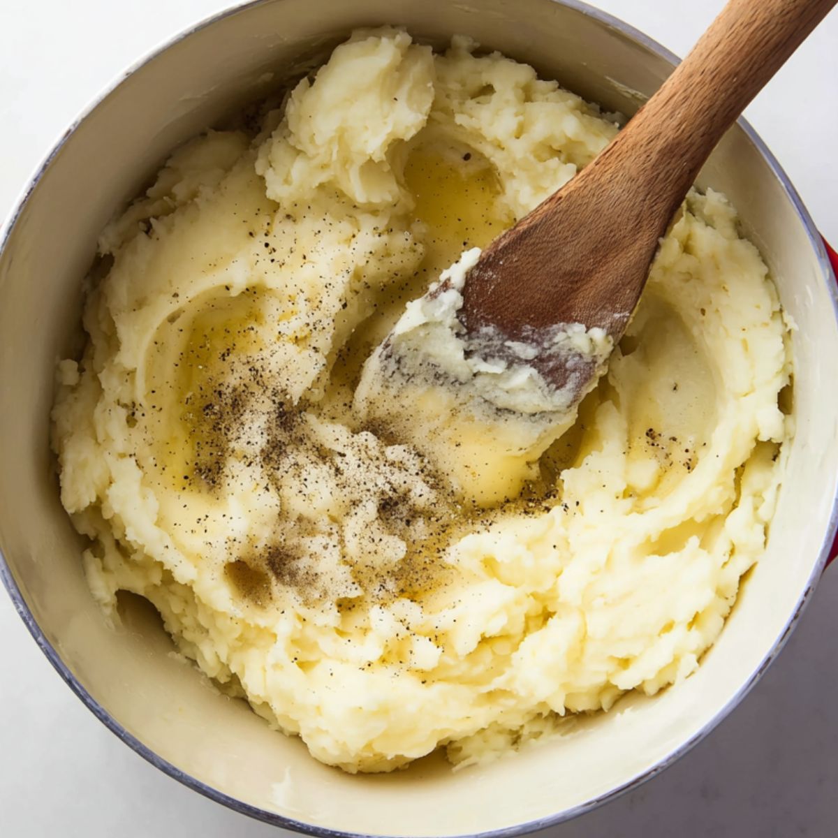 A pot of creamy mashed potatoes being stirred with a wooden spoon, topped with melted butter and freshly ground black pepper.