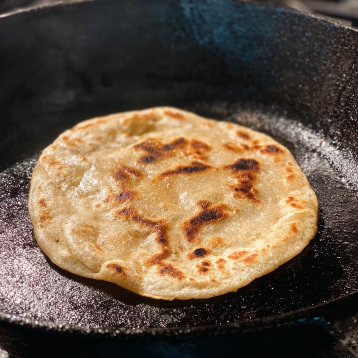 Capture the process of making a perfectly golden, crispy parotta on a cast-iron skillet, with the dough gently browning and puffing up as it cooks. The sizzling sound and the golden crust highlight the skill and care put into making this traditional flatbread