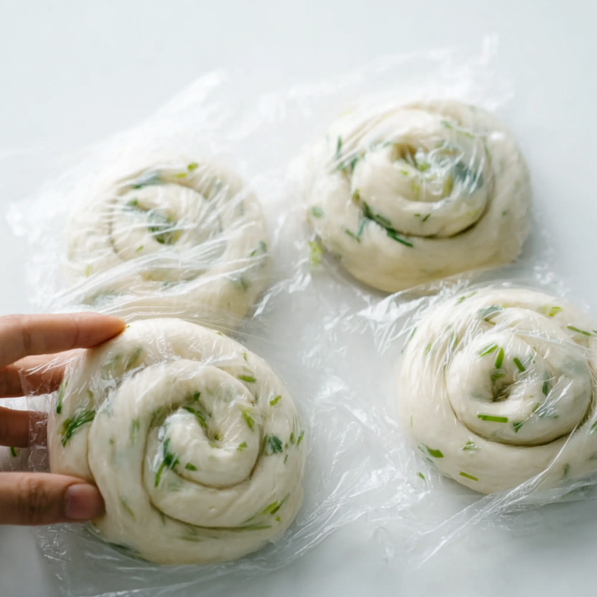 Uncooked scallion pancake dough shaped into spirals and covered with plastic wrap, with green onion pieces visible in the dough.