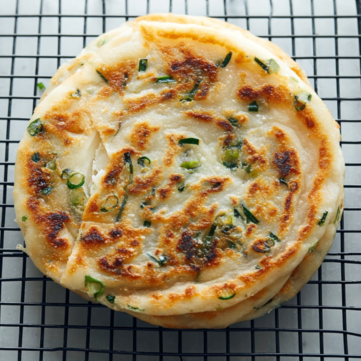 Freshly cooked scallion pancakes cooling on a wire rack, with flaky layers and browned spots visible on the surface.