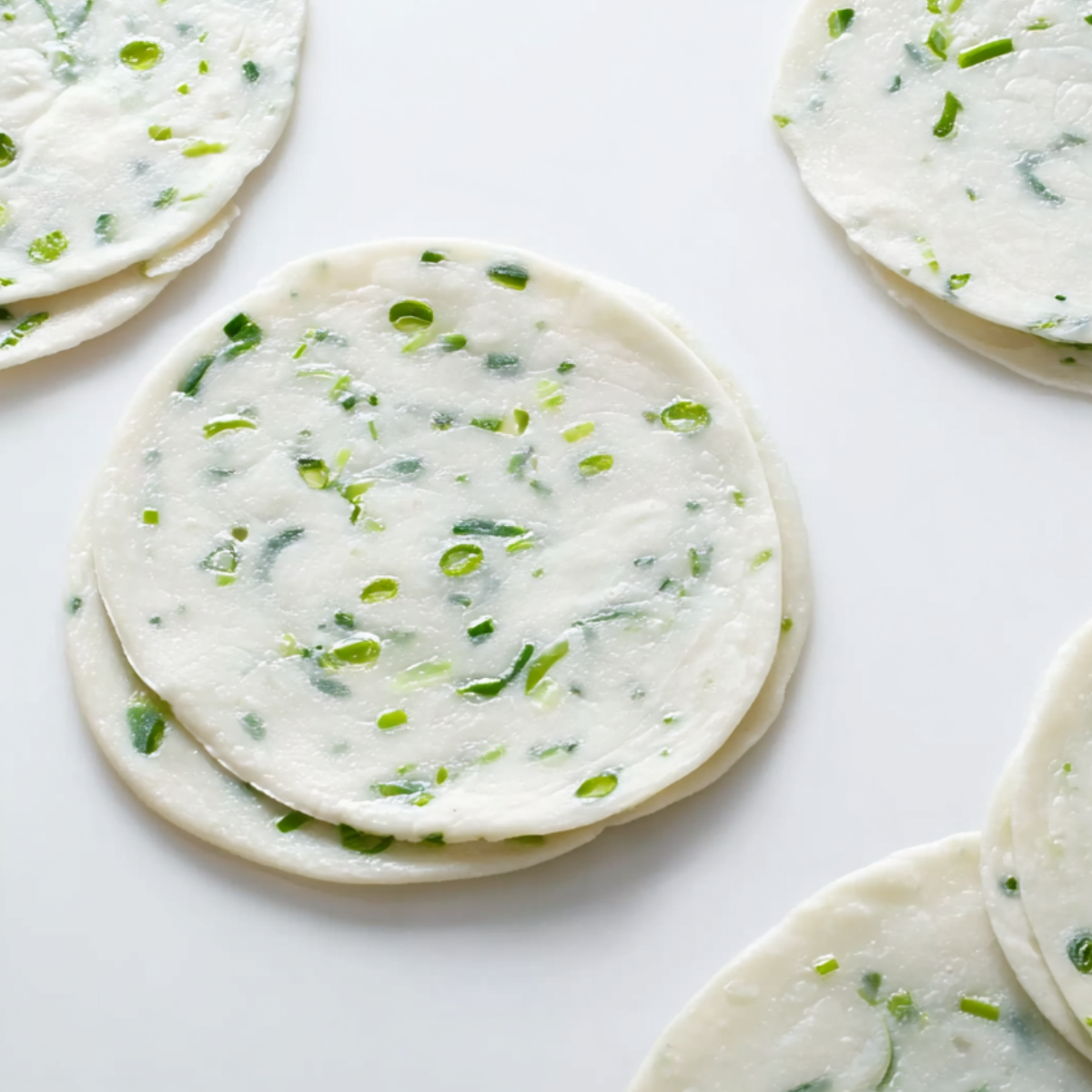 Thin round scallion pancake dough discs laid on a white surface, evenly speckled with chopped green onions.
