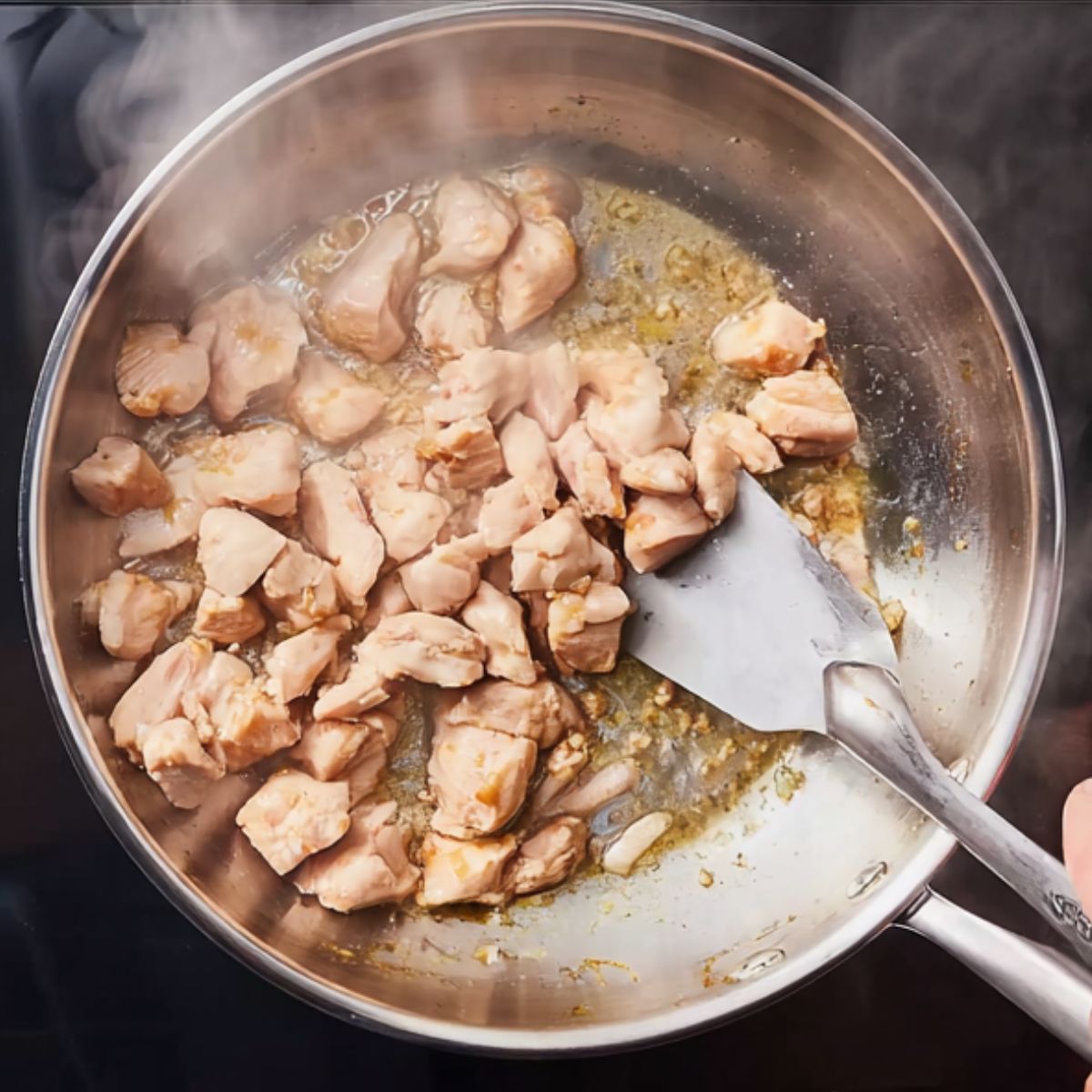 Chicken pieces sautéing in a stainless steel skillet with oil, being stirred with a spatula as steam rises.