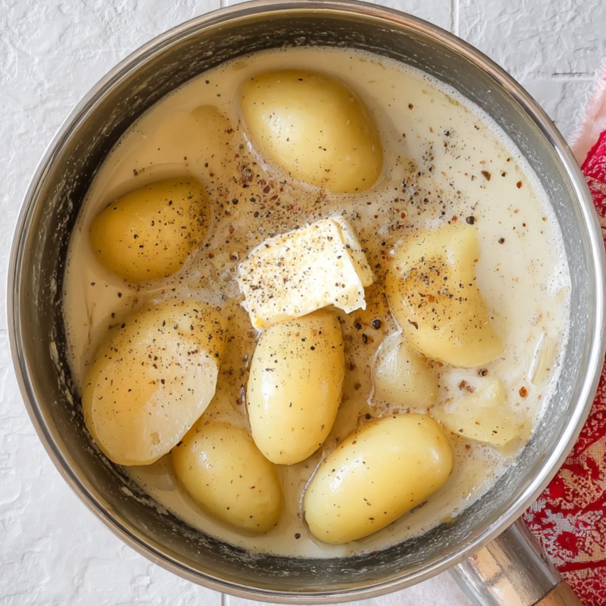 Boiled potatoes simmering in milk with butter and black pepper in a stainless steel saucepan on a white tiled surface.