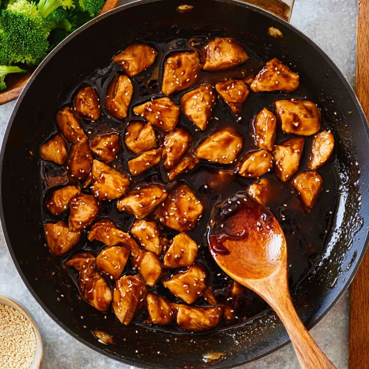 Capture the scene of tender chicken chunks sizzling in a hot pan, coated in a rich, glossy teriyaki sauce. The chicken is perfectly caramelized, with sesame seeds scattered over the sauce, giving it an inviting, flavorful sheen. A wooden spoon stirs the ingredients as they cook, blending the savory sauce with the chicken.