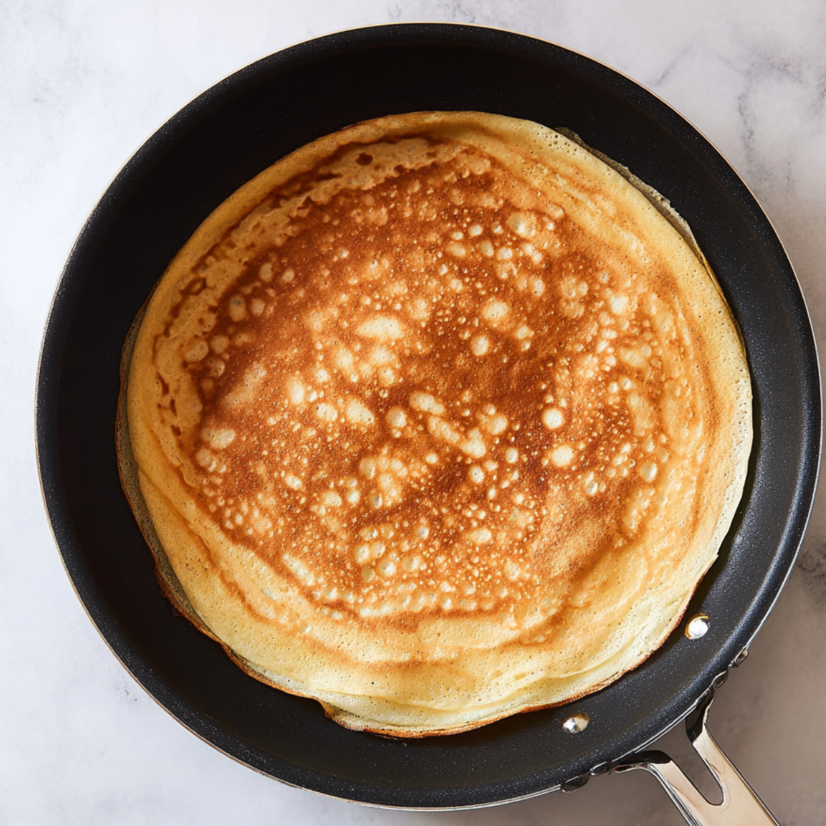 Golden crepe cooking in a black nonstick skillet, photographed from overhead on a light marble surface.