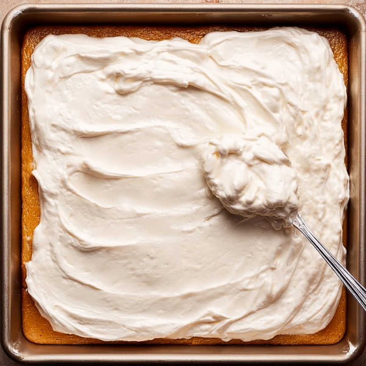 A rectangular pan of cake topped with a smooth layer of whipped cream, with a spoon spreading the cream evenly over the surface.