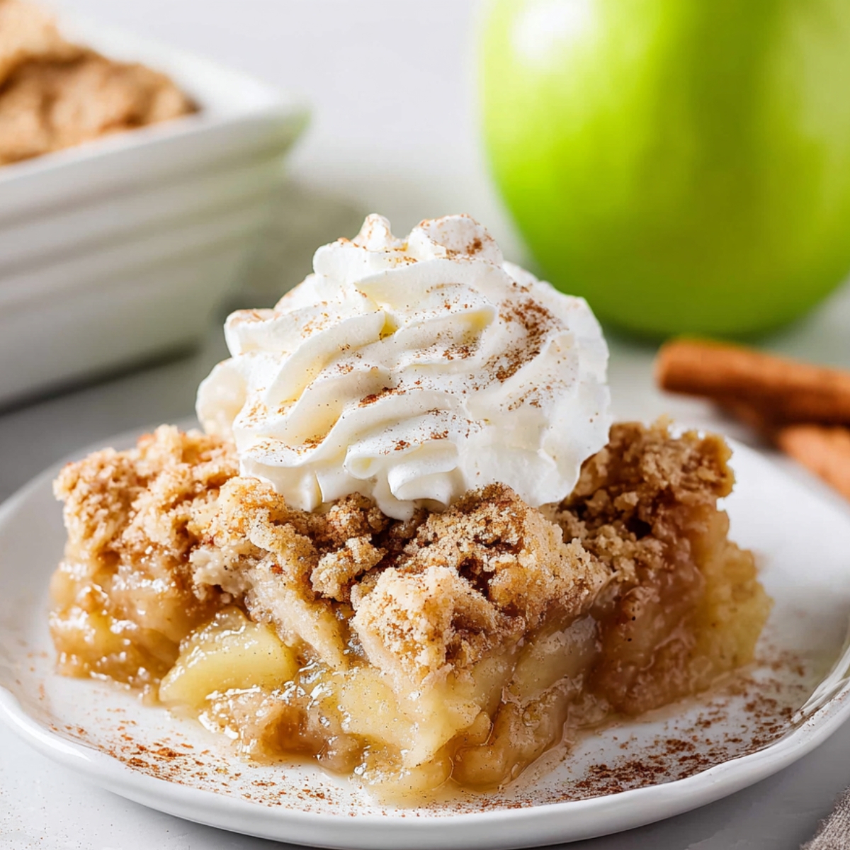 Apple Dump Cake recipe
Slice of apple dump cake on a white plate topped with whipped cream and cinnamon, showing warm apple filling and crumb topping with cinnamon sticks in the background.