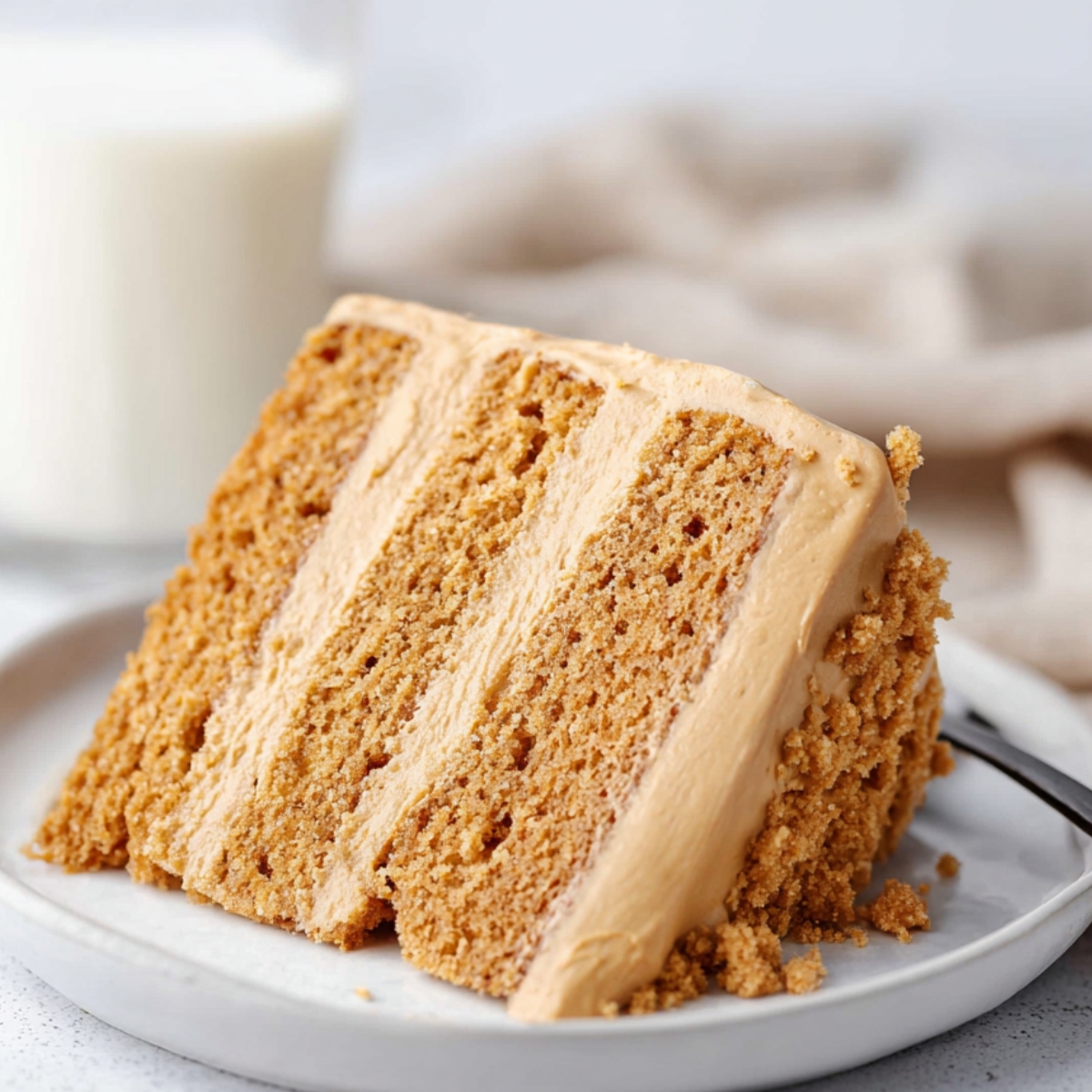 Biscoff Cake Recipe Slice of homemade Biscoff layer cake with creamy cookie butter frosting on a white plate, served with a glass of milk in the background.