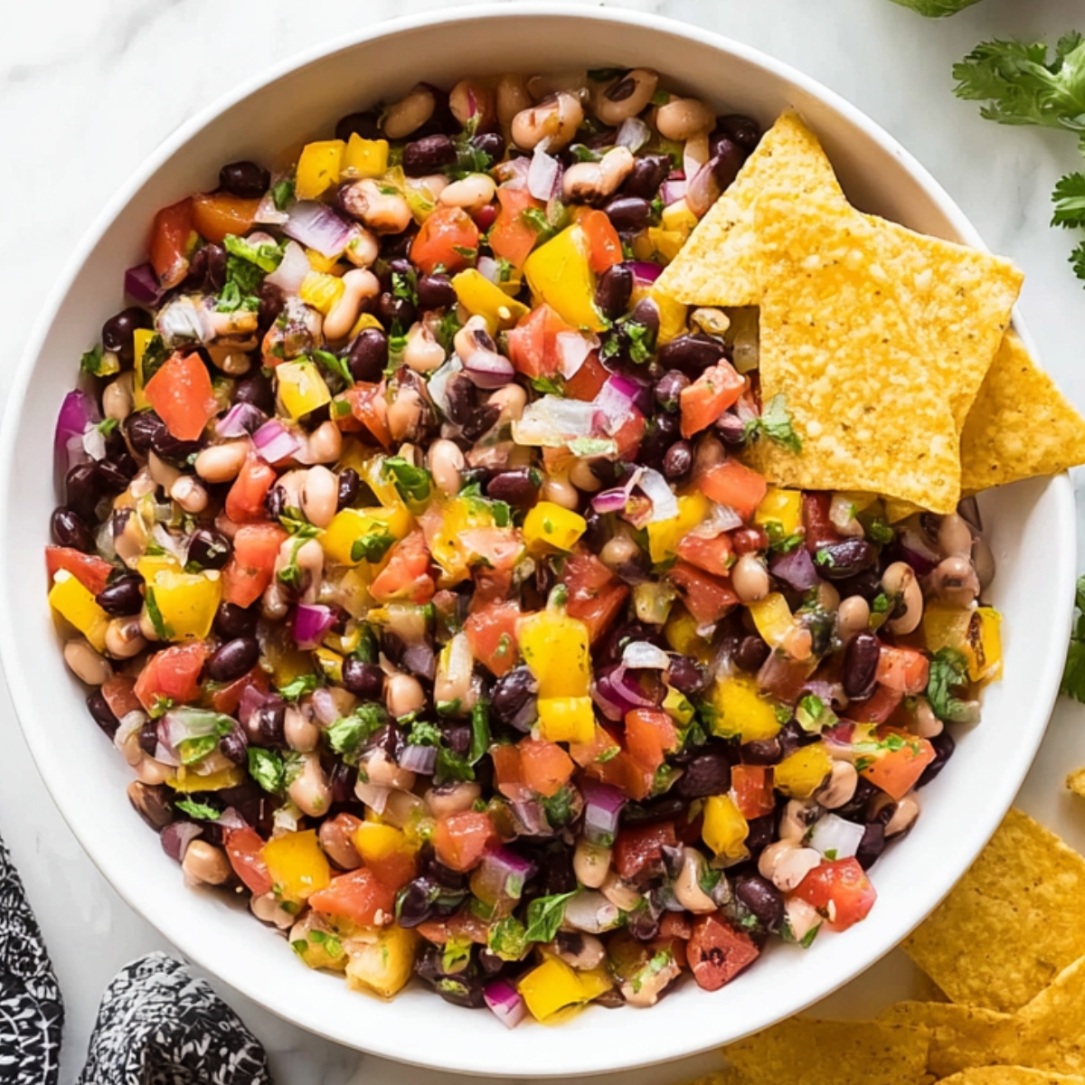 Cowboy Caviar Overhead view of a white bowl filled with colorful cowboy caviar made with black beans, black-eyed peas, diced tomatoes, yellow bell peppers, red onion, and cilantro, topped with tortilla chips on a marble surface.