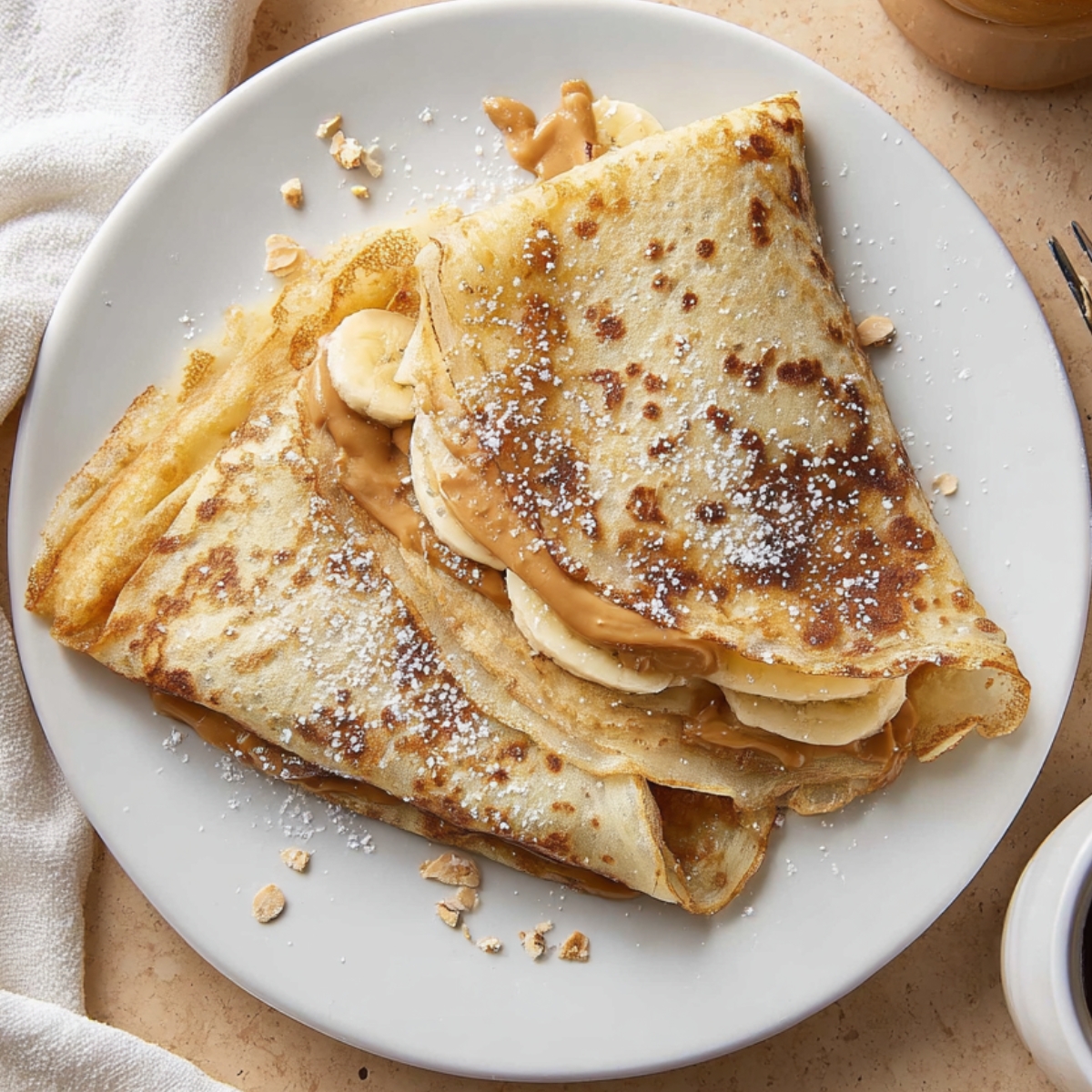 easy homemade crepes Overhead breakfast scene of folded crepes filled with banana and peanut butter on a white ceramic plate. The crepes are lightly golden with brown spots and dusted with powdered sugar. Peanut butter is slightly melted and visible inside. A fork sits on the right, a white cloth napkin on the left, a cup of black coffee in the top corner, and a jar of peanut butter in the background. Warm soft lighting, cozy morning atmosphere, realistic food photography, high detail, 4k resolution.